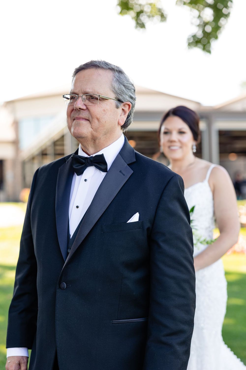 Father of the bride in tuxedo stands outdoors as smiling bride approaches for first look reveal.