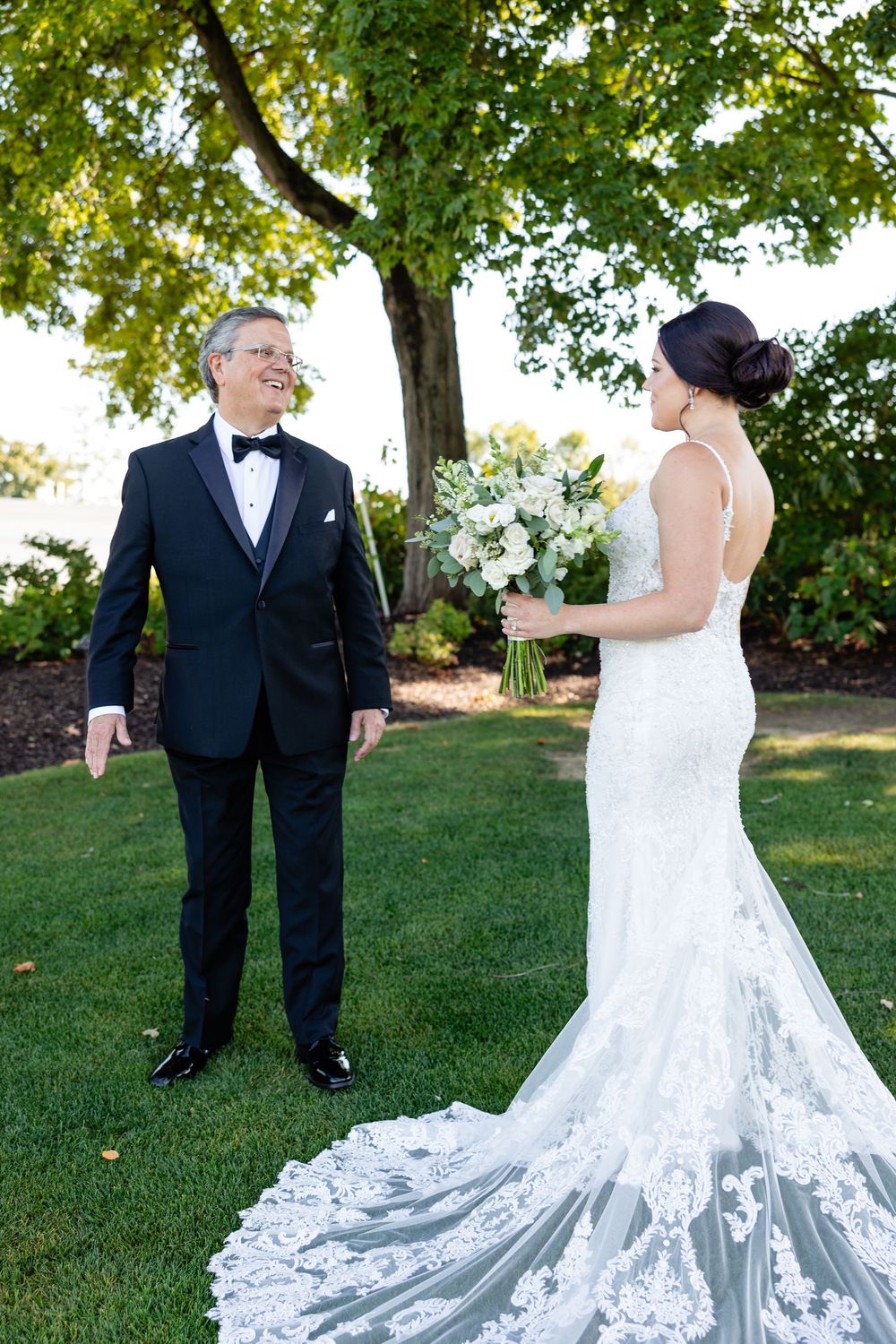 Bride in lace gown shows first look to father in tuxedo on green lawn under trees.