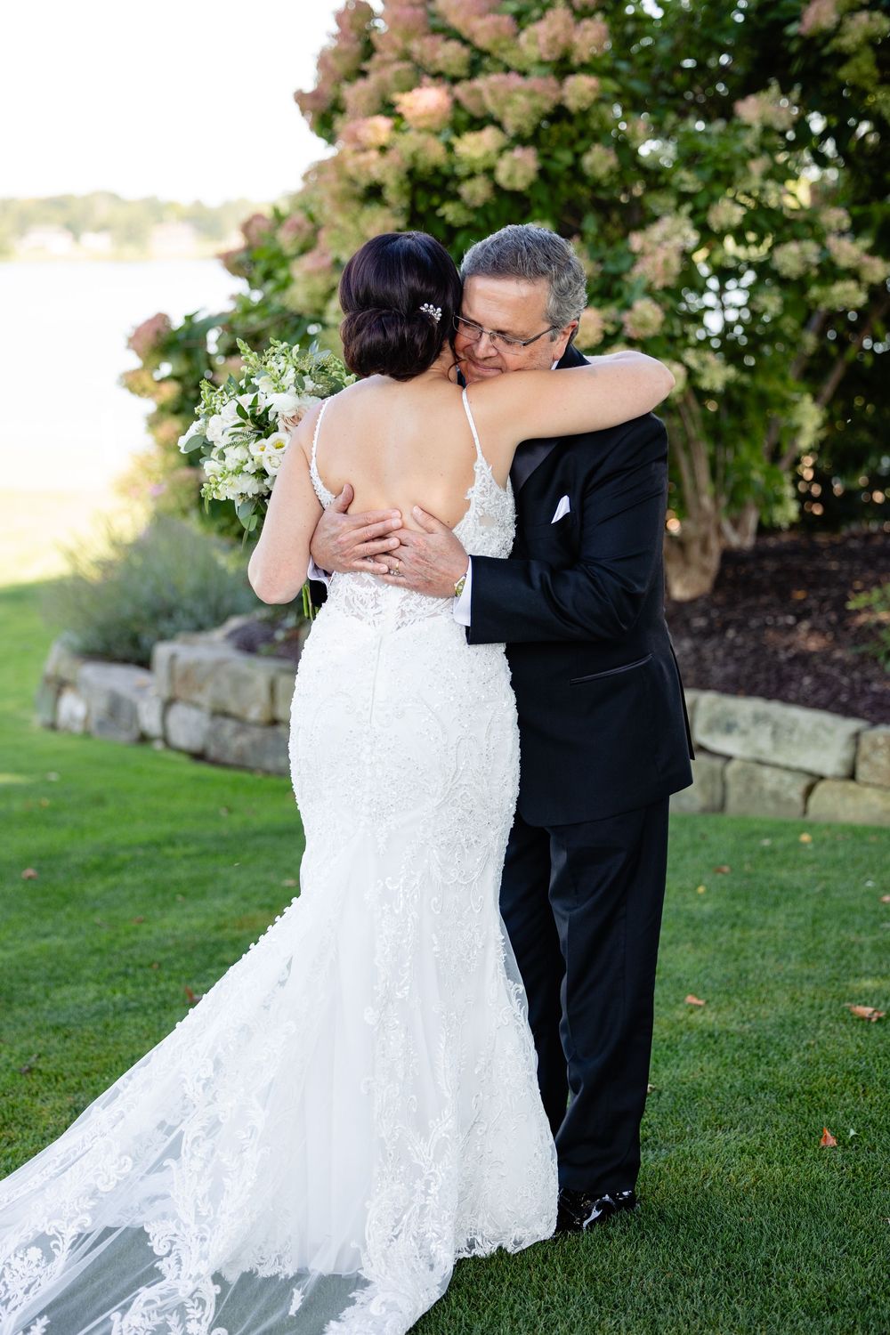 Bride in lace mermaid gown embraces older man in tuxedo during emotional first look at outdoor wedding.