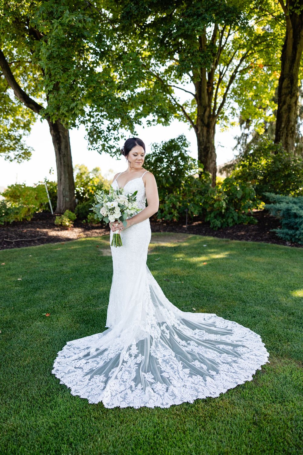 Bride in lace mermaid wedding gown with long train holds white bouquet outdoors on green lawn beneath trees.