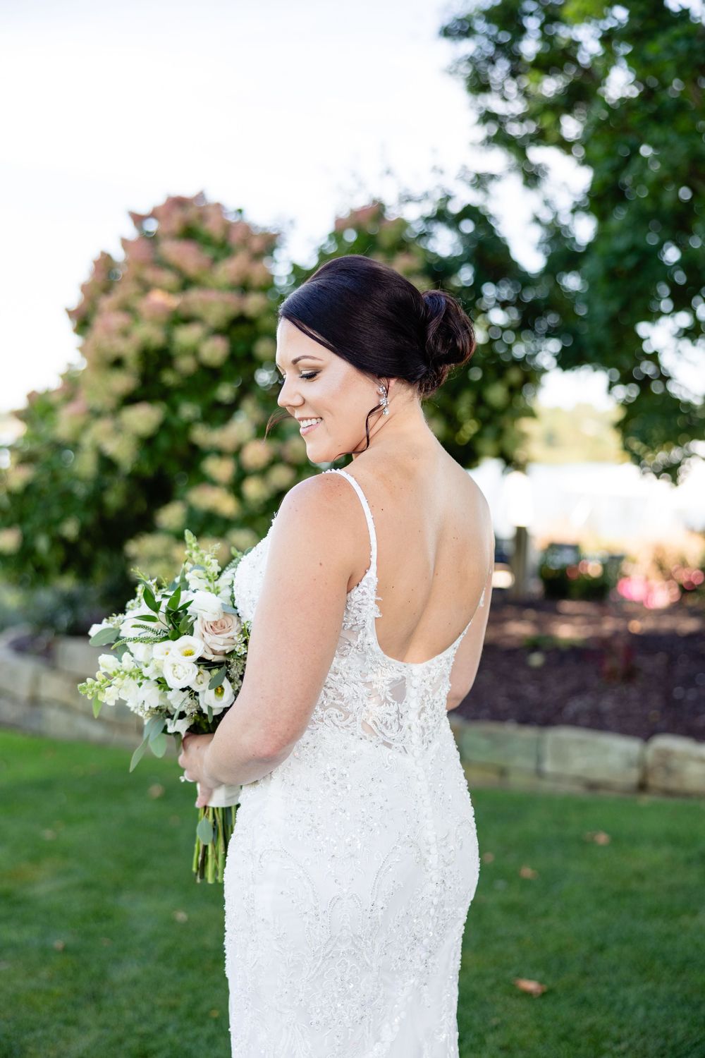 Bride in lace backless wedding gown holds white bouquet, smiling over shoulder in garden.
