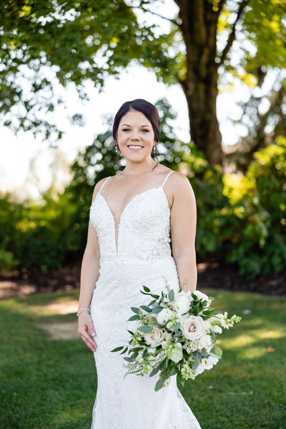 Smiling bride in beaded spaghetti strap gown holding white and green bouquet outdoors.