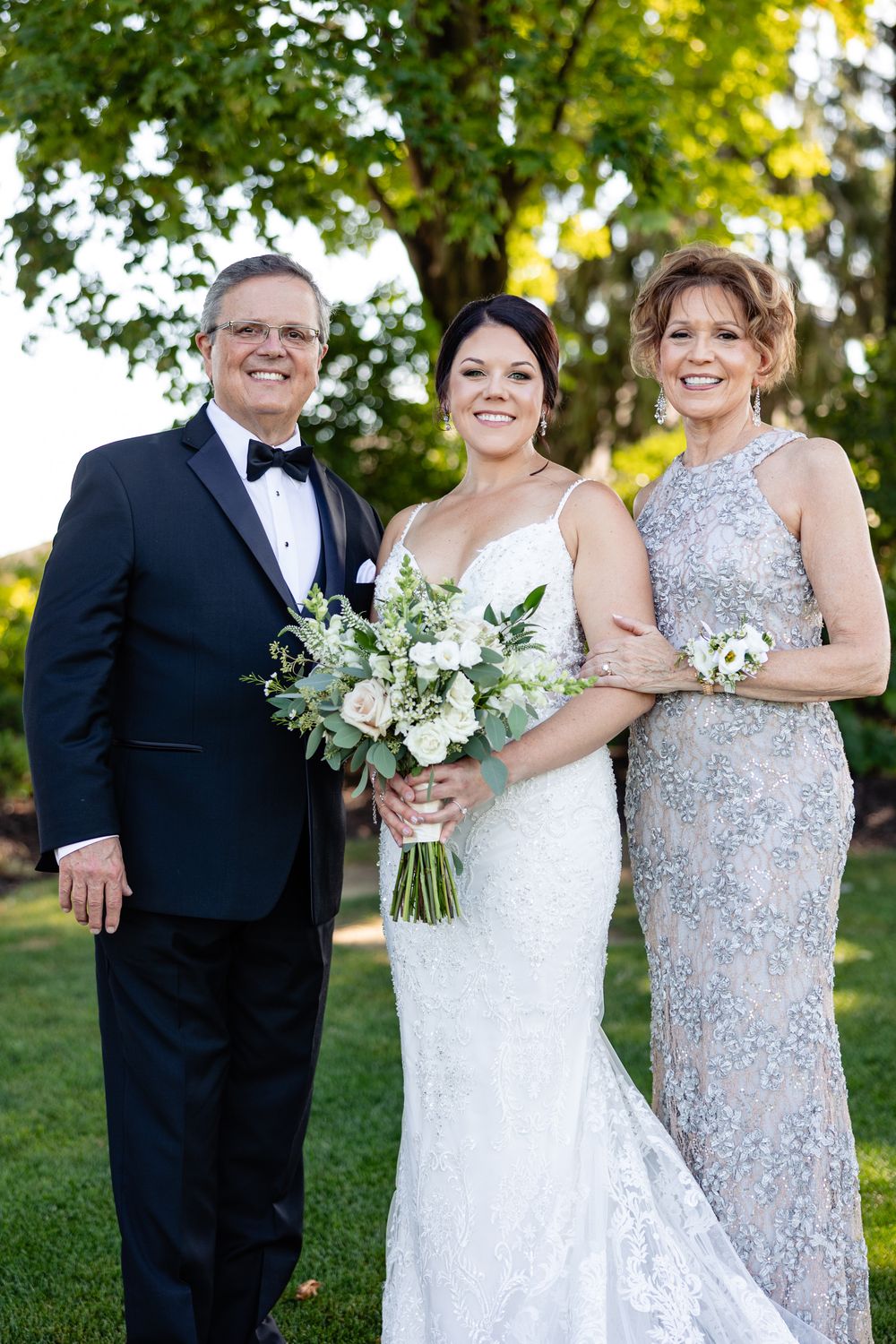 Bride in lace gown poses with parents, father in tuxedo and mother in silver gown, outdoors.