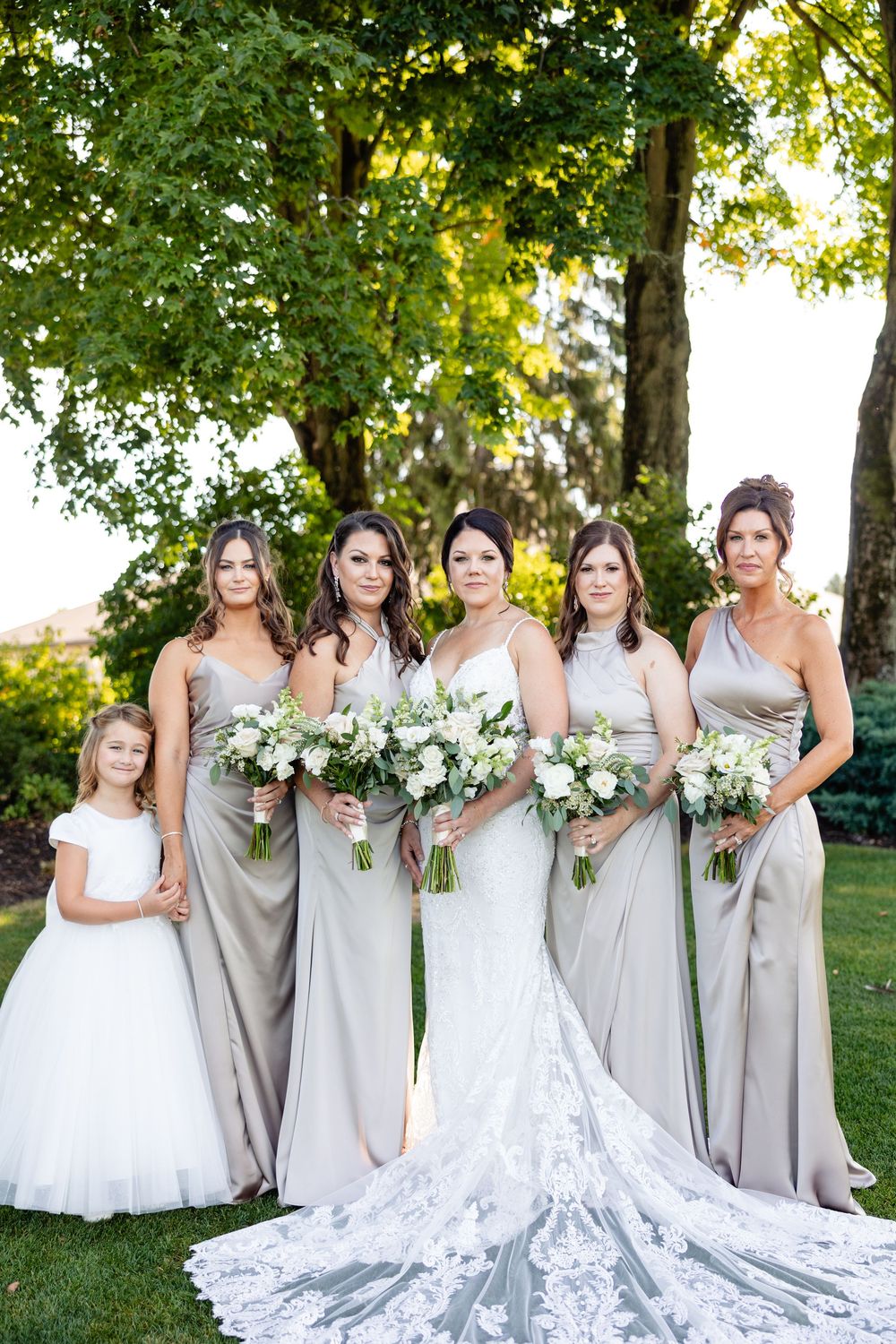Bride in lace gown with bridesmaids in taupe satin dresses and flower girl holding white bouquets outdoors.