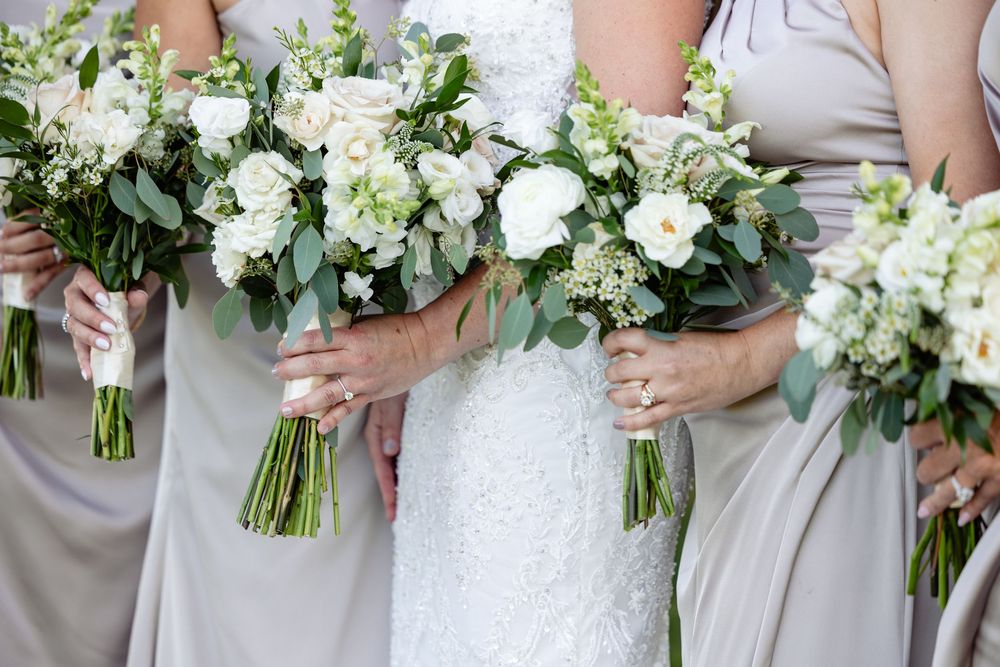 Bride and bridesmaids holding white rose and greenery bouquets, wearing white lace and gray dresses.
