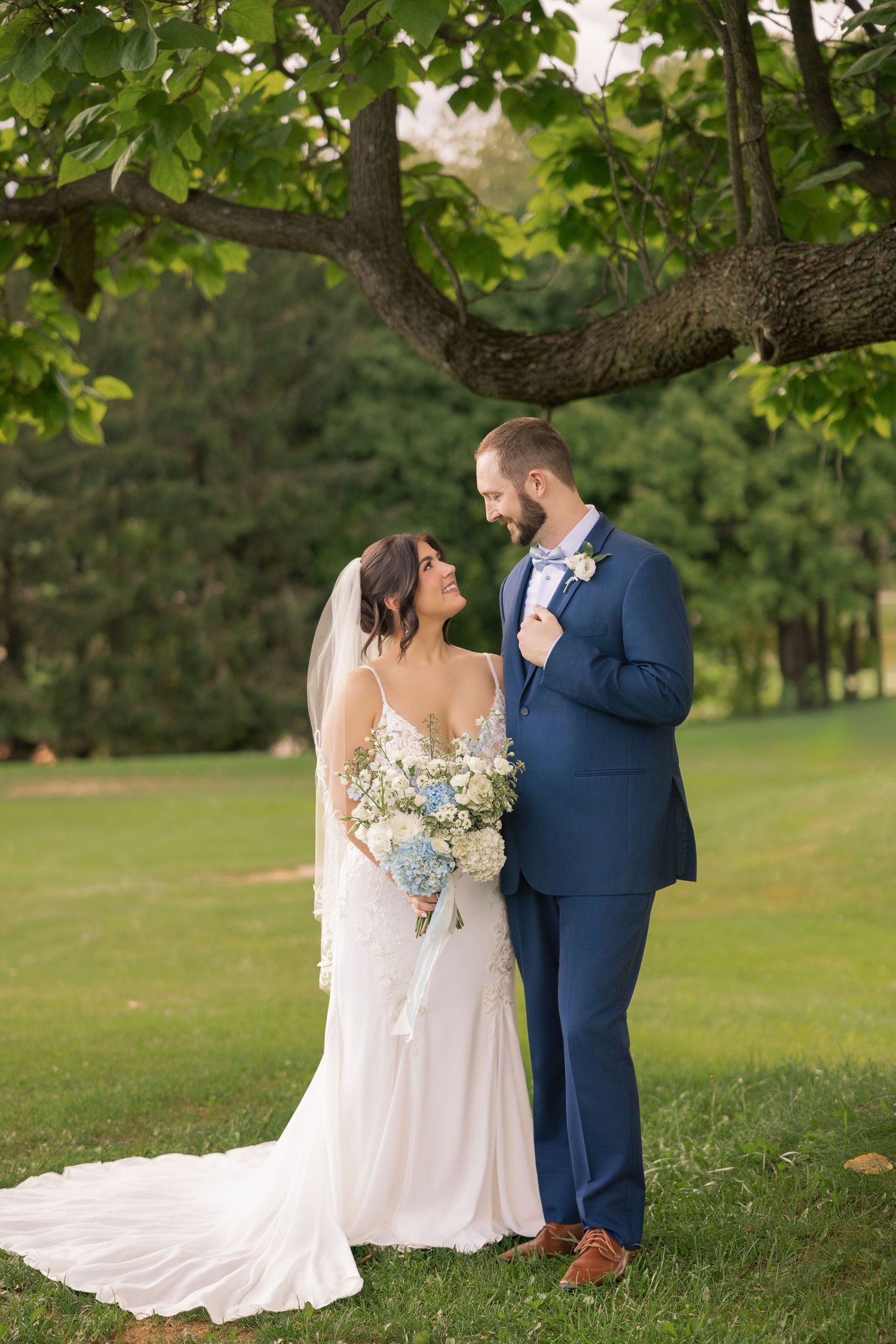 Bride and groom share a romantic moment under a tree, she in a white gown with veil, he in a navy suit.