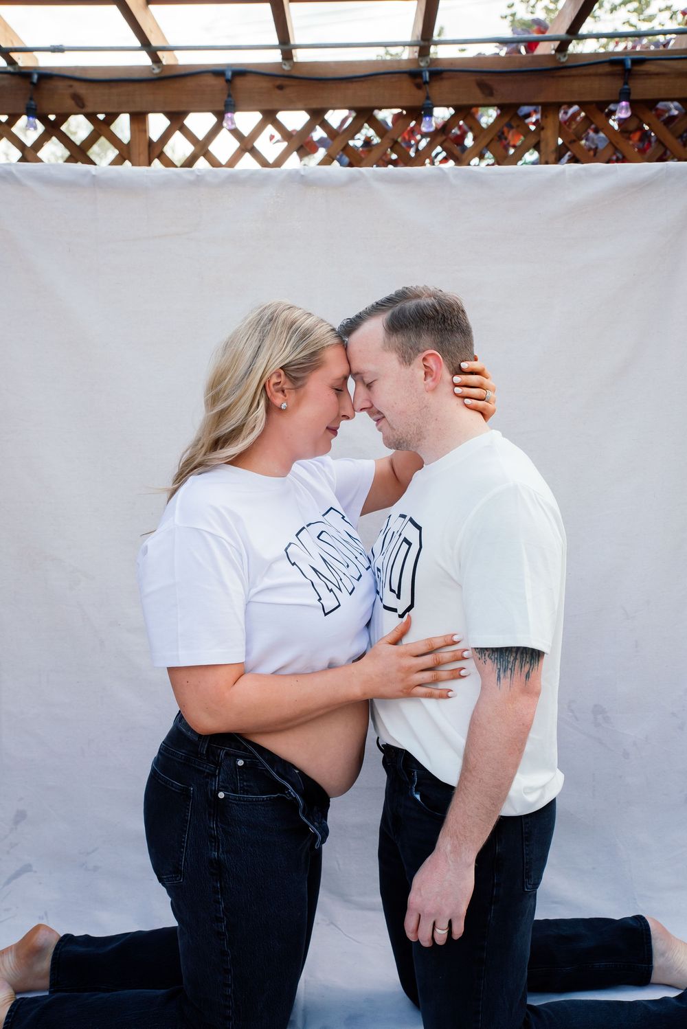 Pregnant couple in white shirts touching foreheads tenderly outdoors against white backdrop.