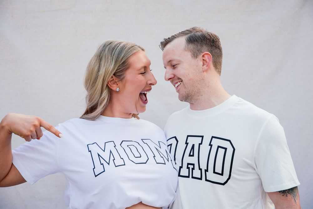 Couple wearing matching white Mom and Dad graphic t-shirts laughing together against a white background.