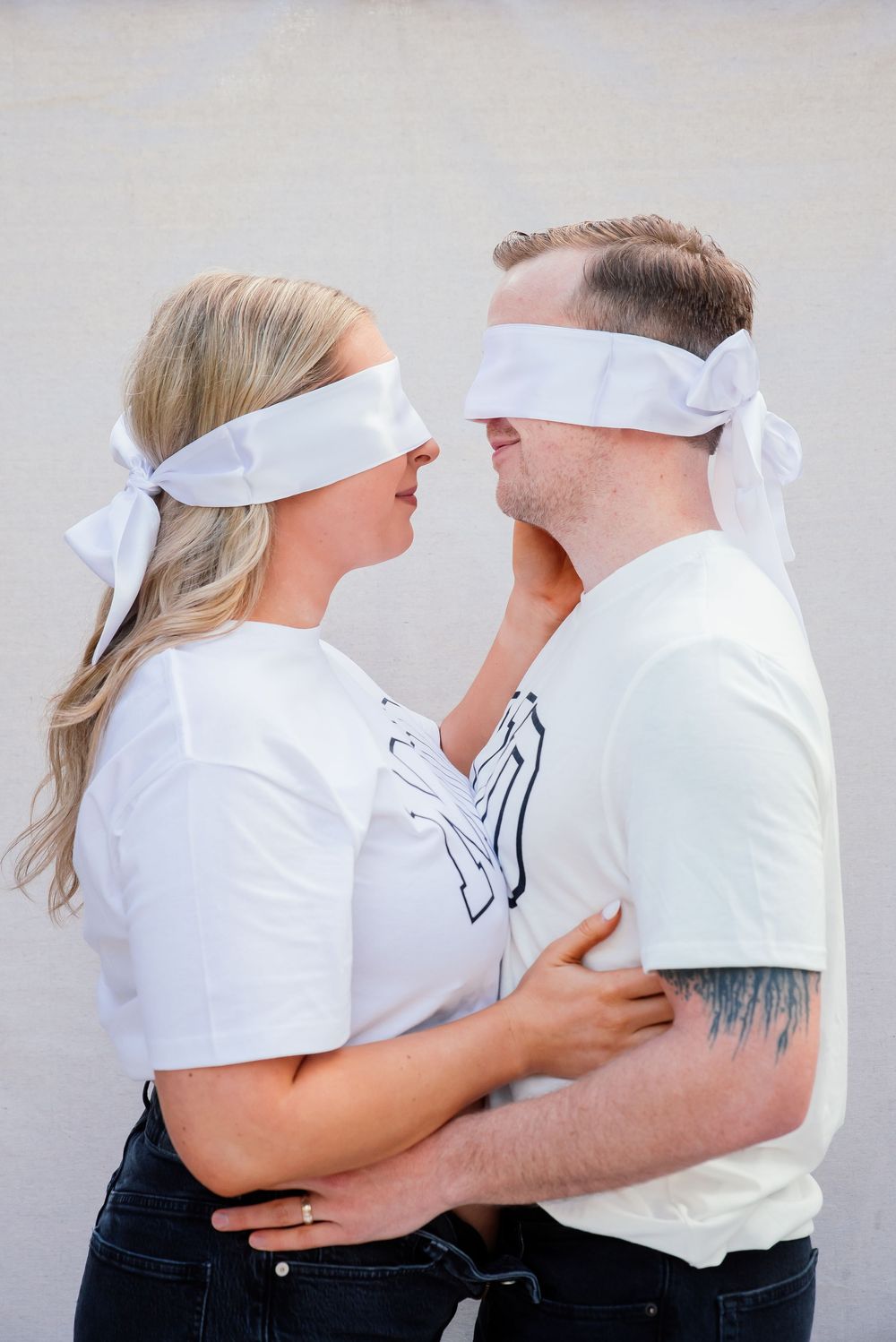 Couple wearing white blindfolds embracing each other against a light gray wall in matching white t-shirts.