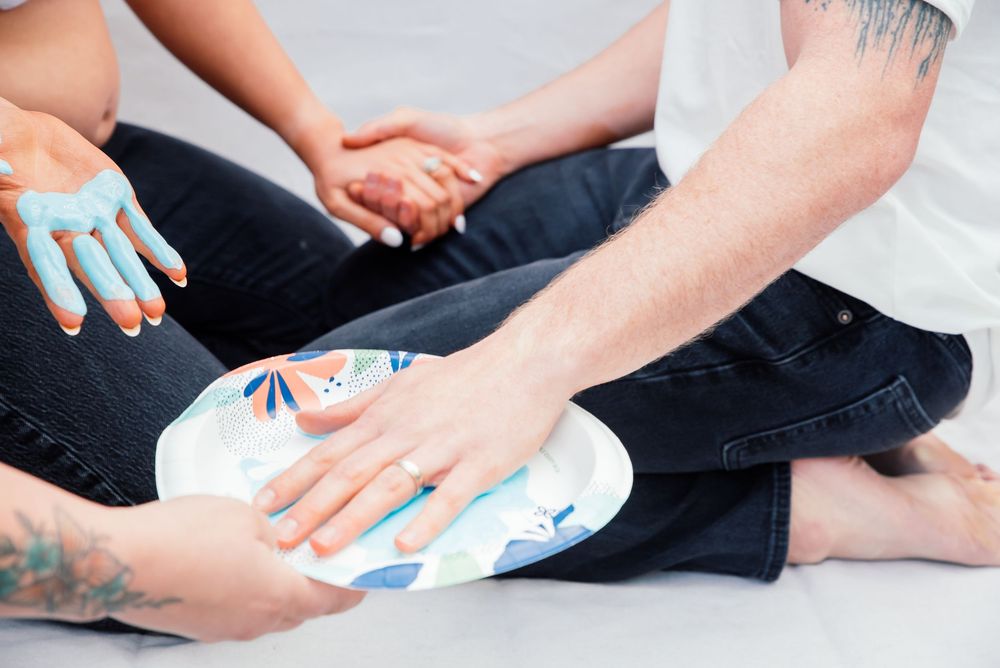 Two people with tattoos sit together, one holding a decorative paper plate with painted hands nearby.