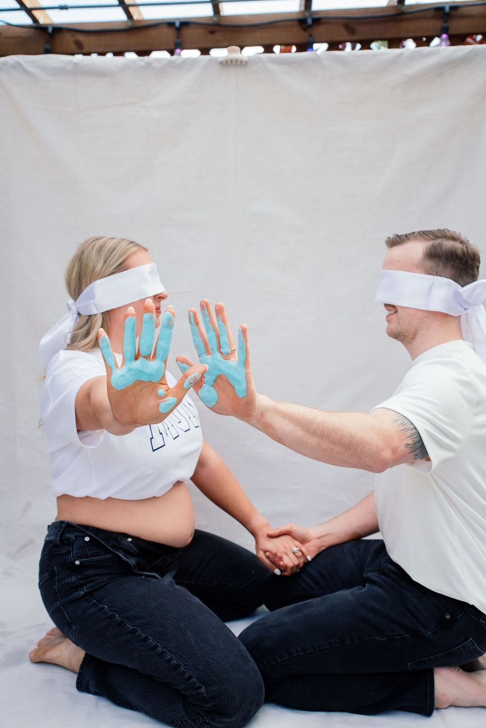 Blindfolded couple sitting cross-legged during gender reveal, pregnant woman showing blue paint-covered hands, indicating a baby boy.