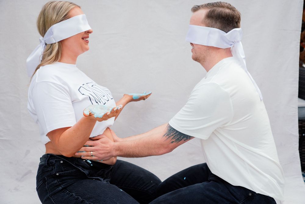 Two blindfolded people in white shirts interact playfully during a gender reveal party, with blue powder visible.