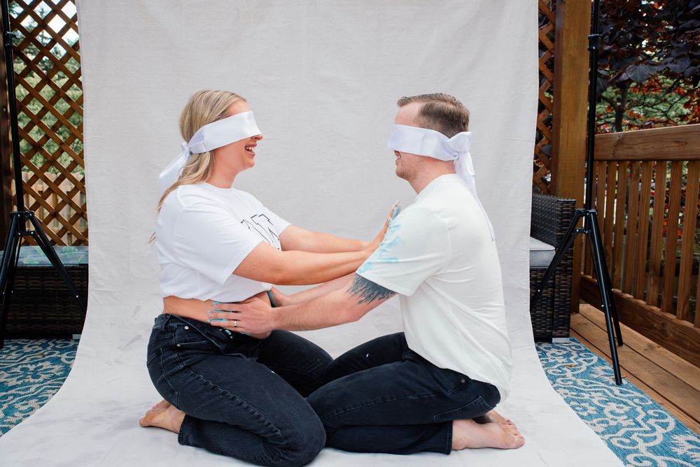 Blindfolded couple sitting face-to-face on a patterned rug, touching each other's arms in a trust exercise outdoors.