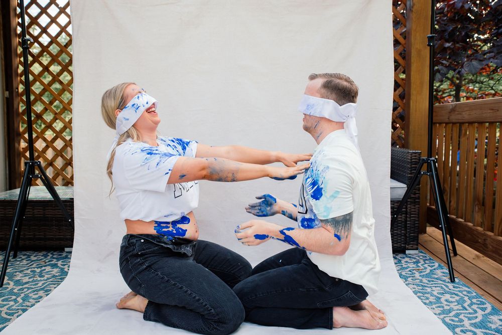 Blindfolded couple playfully smearing blue paint on each other while sitting on a white backdrop outdoors.