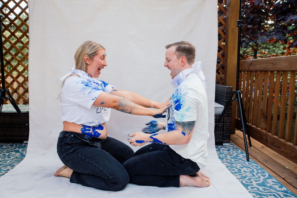 Couple sitting on white sheet laughing while smearing blue paint on each other during a fun gender reveal photo shoot.