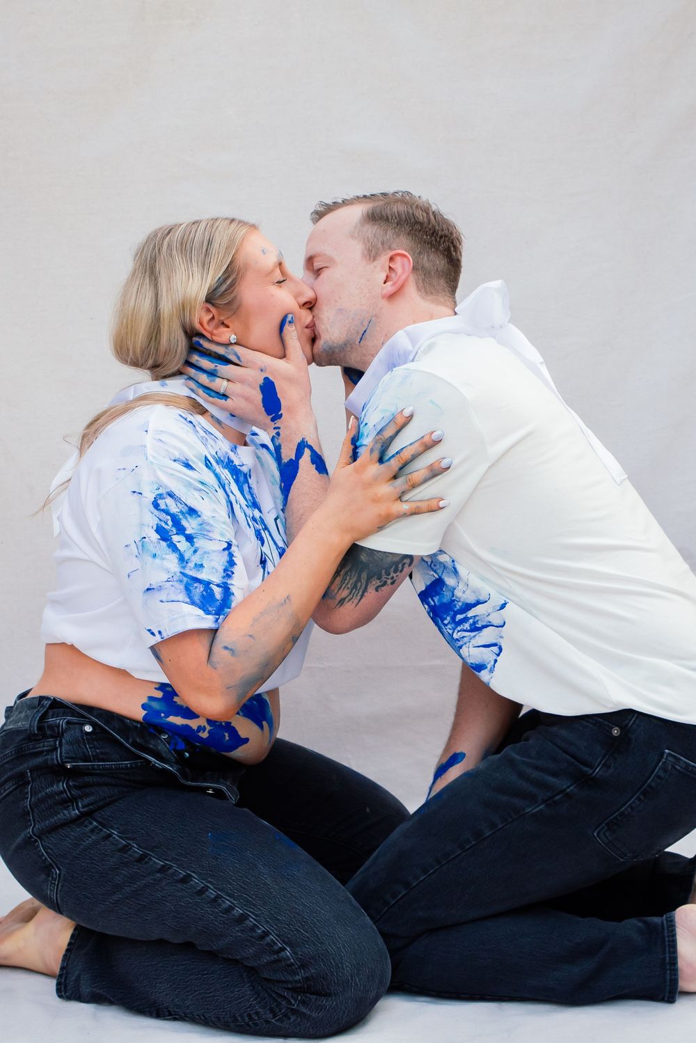 Couple kissing while covered in blue paint, sitting on floor in white shirts and dark jeans.