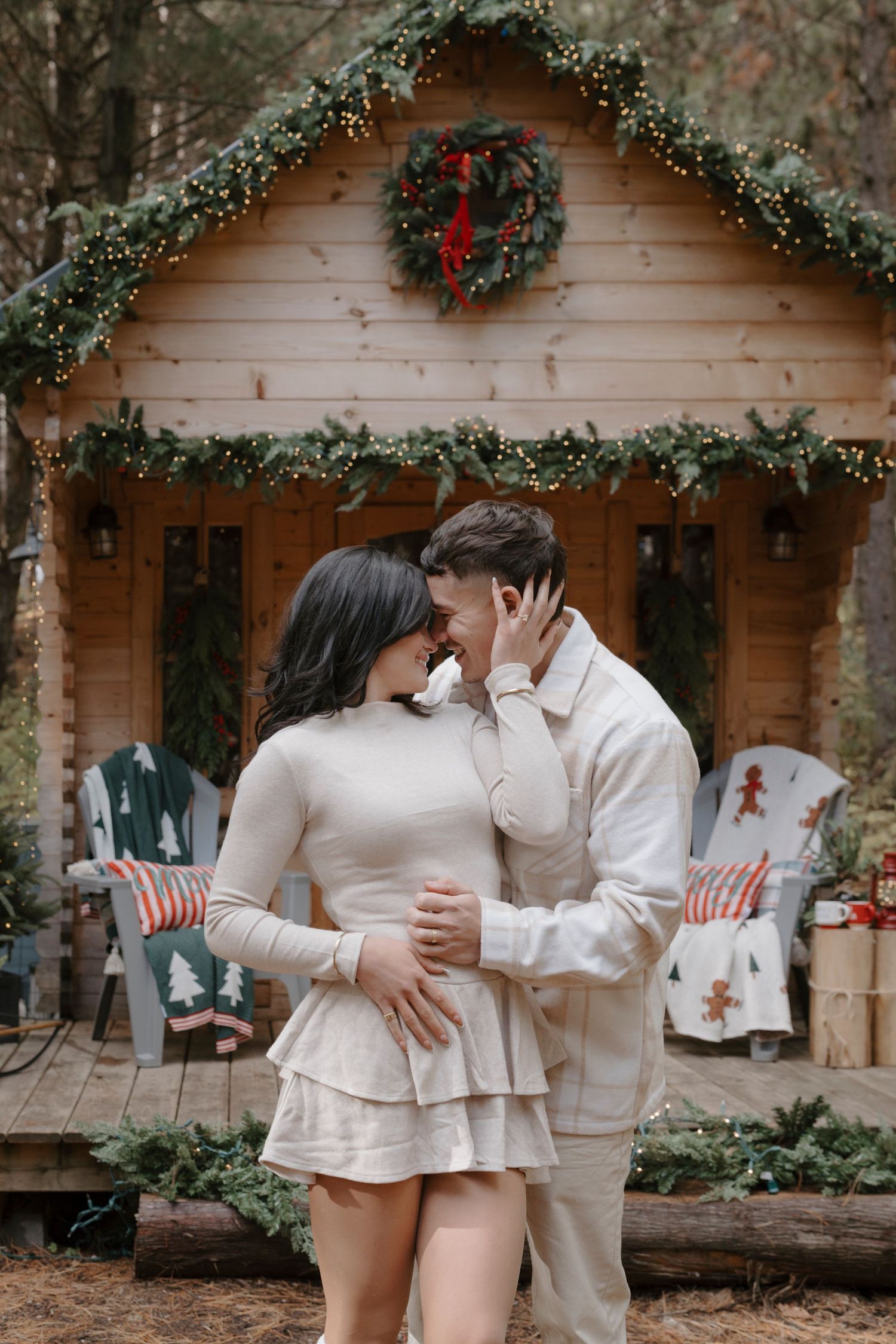 Couple embracing romantically in front of a Christmas-decorated wooden cabin with wreaths and fairy lights.