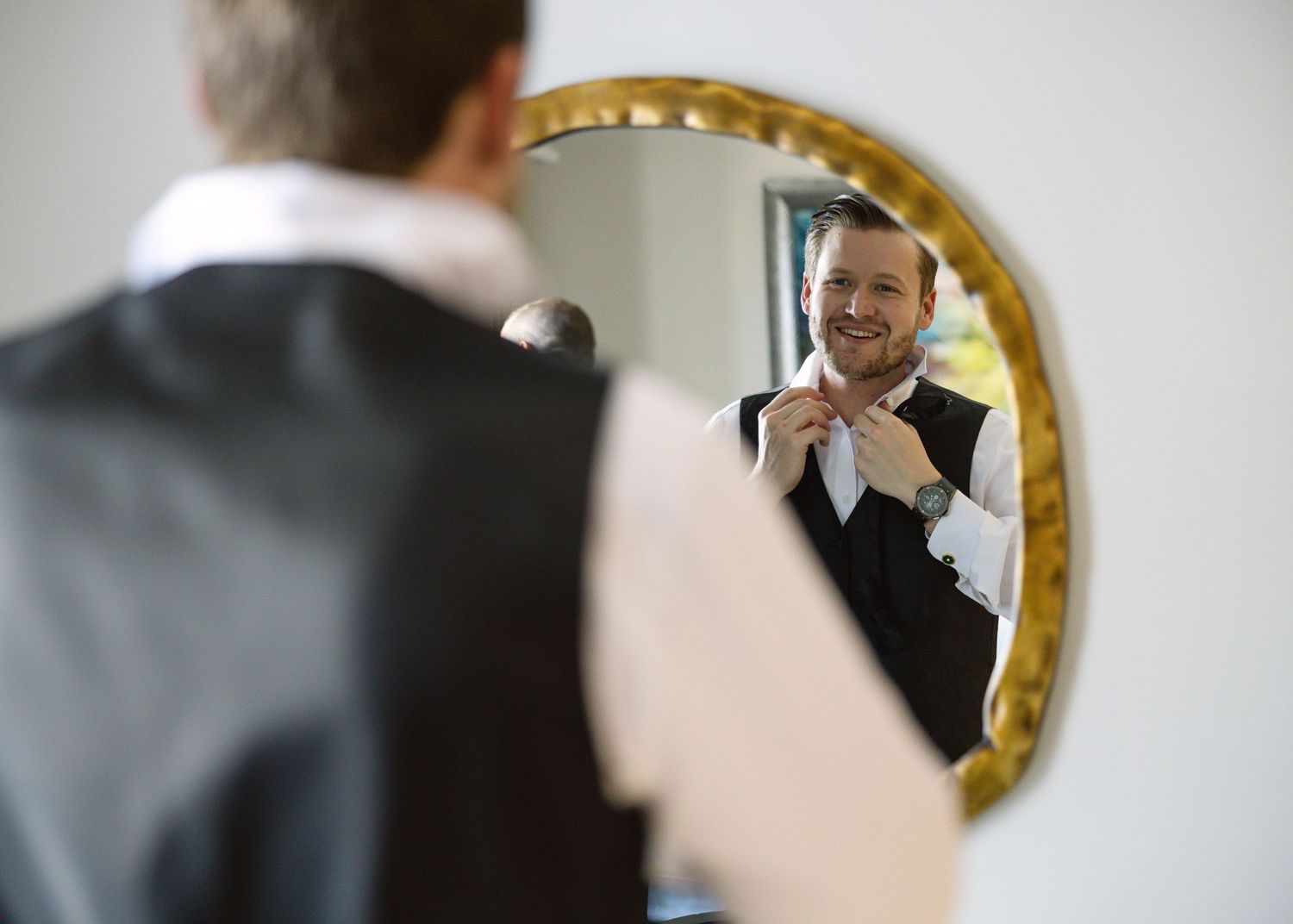 Smiling groom adjusting his tie in an ornate gold-framed mirror while dressed in a black vest and white shirt.