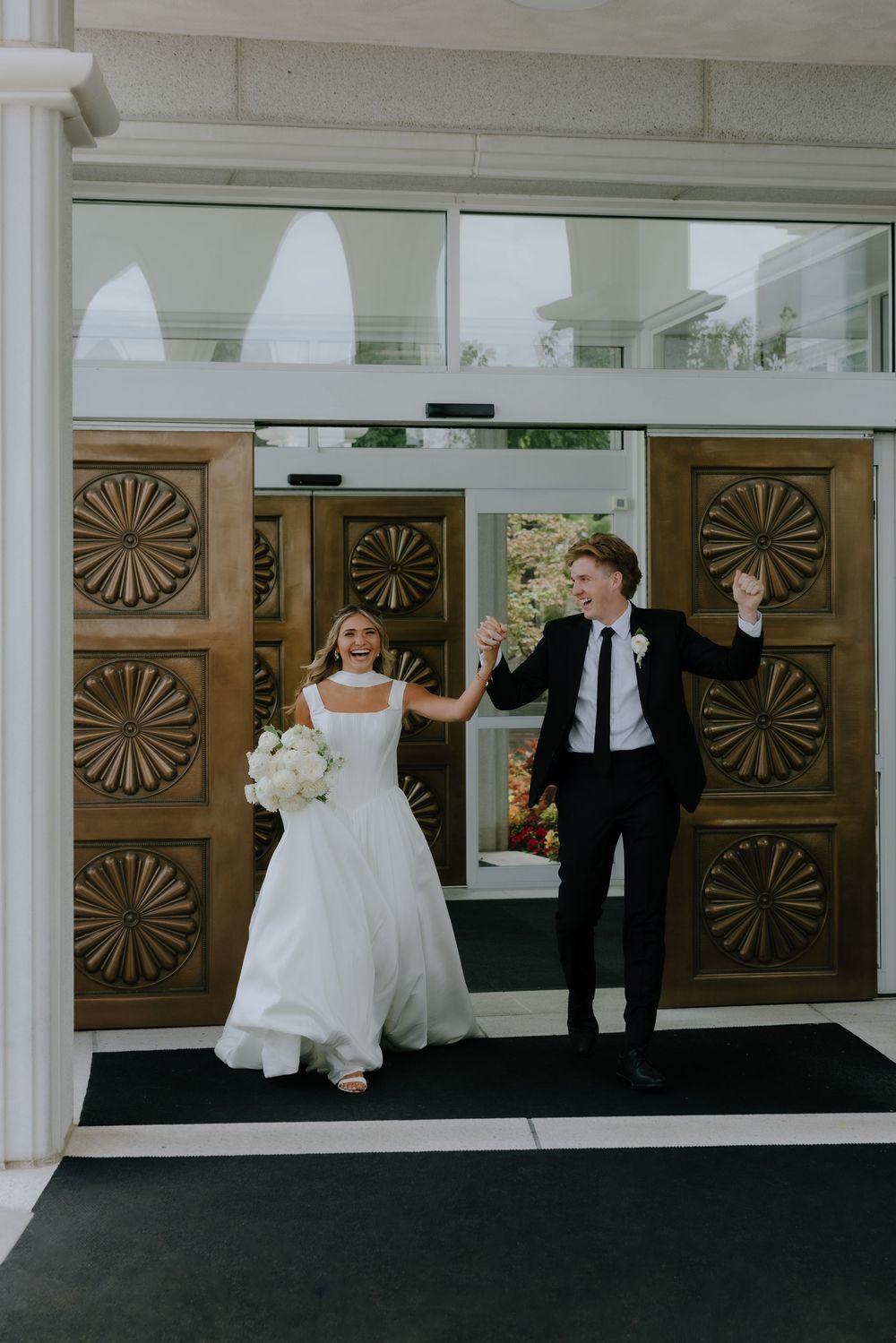 Joyful bride and groom exiting through ornate wooden doors, holding hands and celebrating their wedding day.