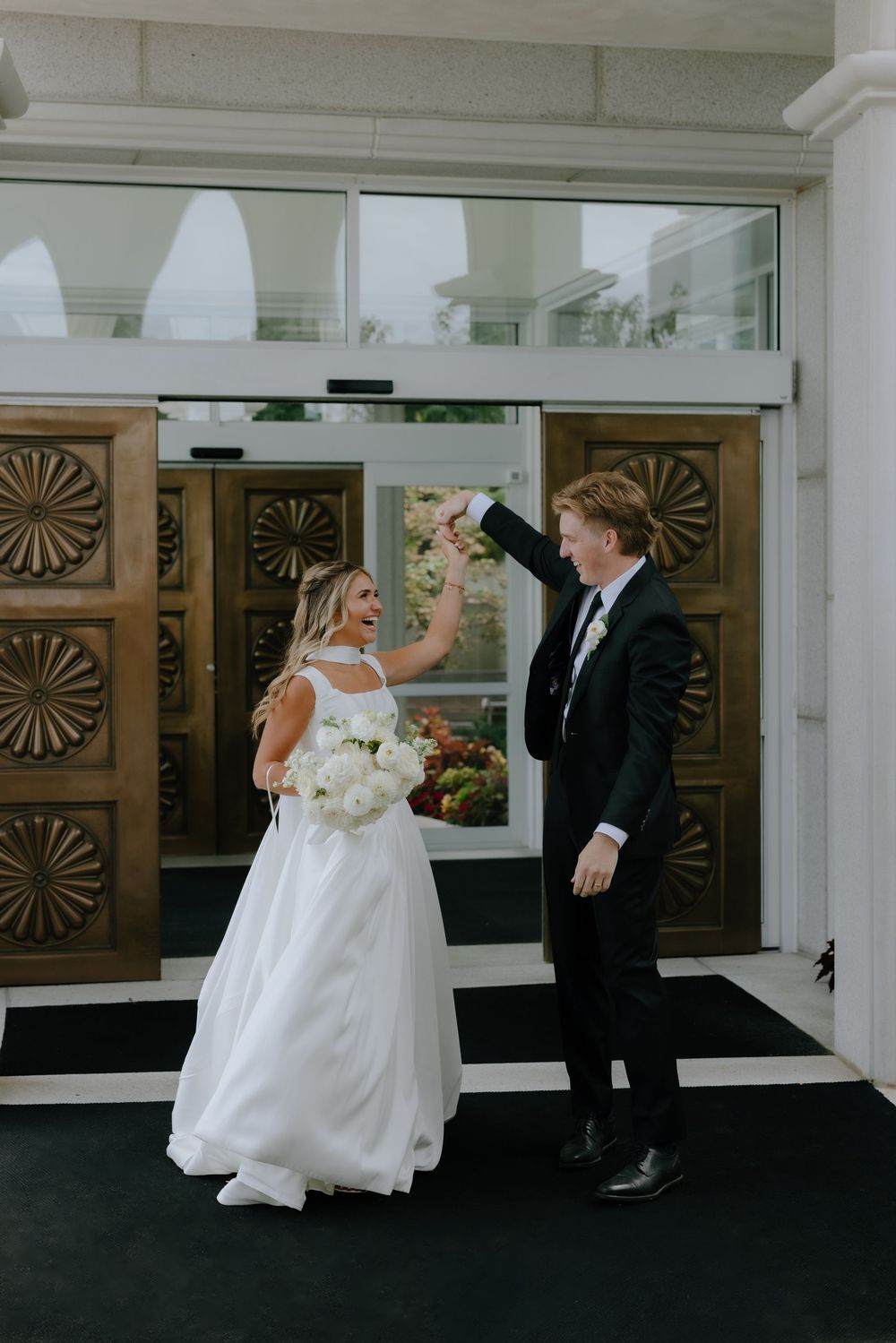 Bride and groom celebrate exiting church doors, bride holding white bouquet while groom raises her hand triumphantly.