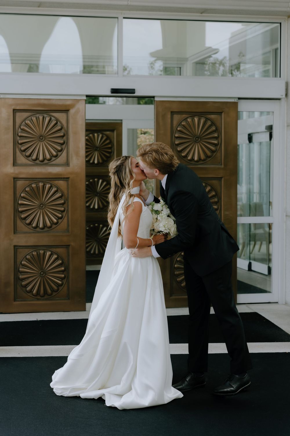 Bride and groom share a first kiss in front of ornate bronze doors on their wedding day.