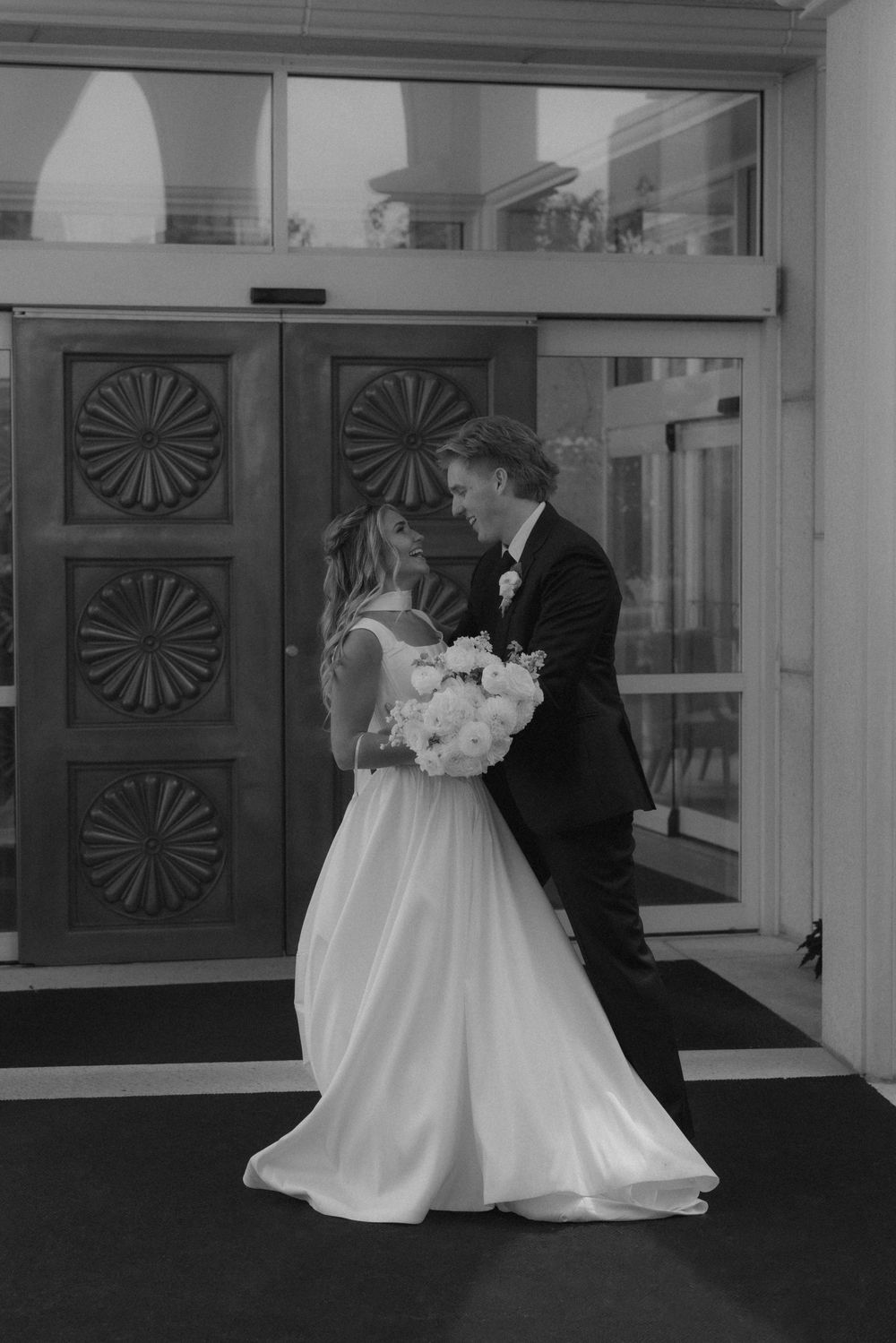 Black and white photo of bride and groom embracing before ornate wooden doors at their wedding venue.