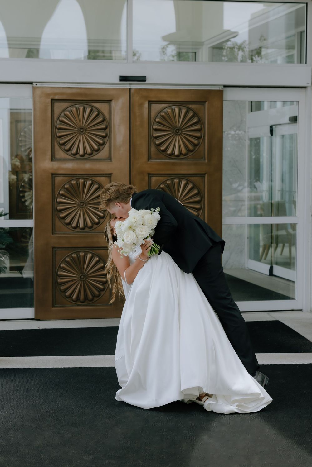 Groom dips bride for a kiss in front of ornate wooden doors, bride holding white floral bouquet.