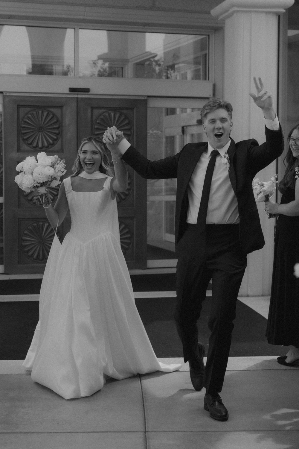 Joyful newlyweds celebrate exit from church doors in black and white, raising hands with bouquet.