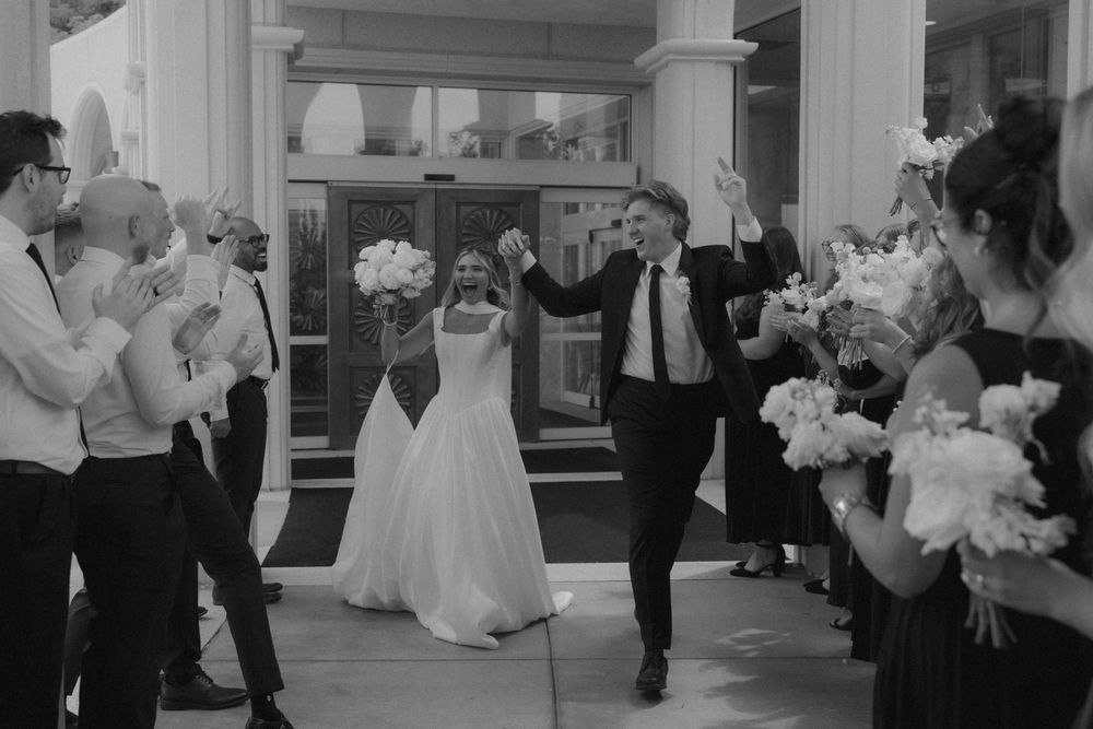 Black and white photo of bride and groom exiting wedding venue through cheering guests.