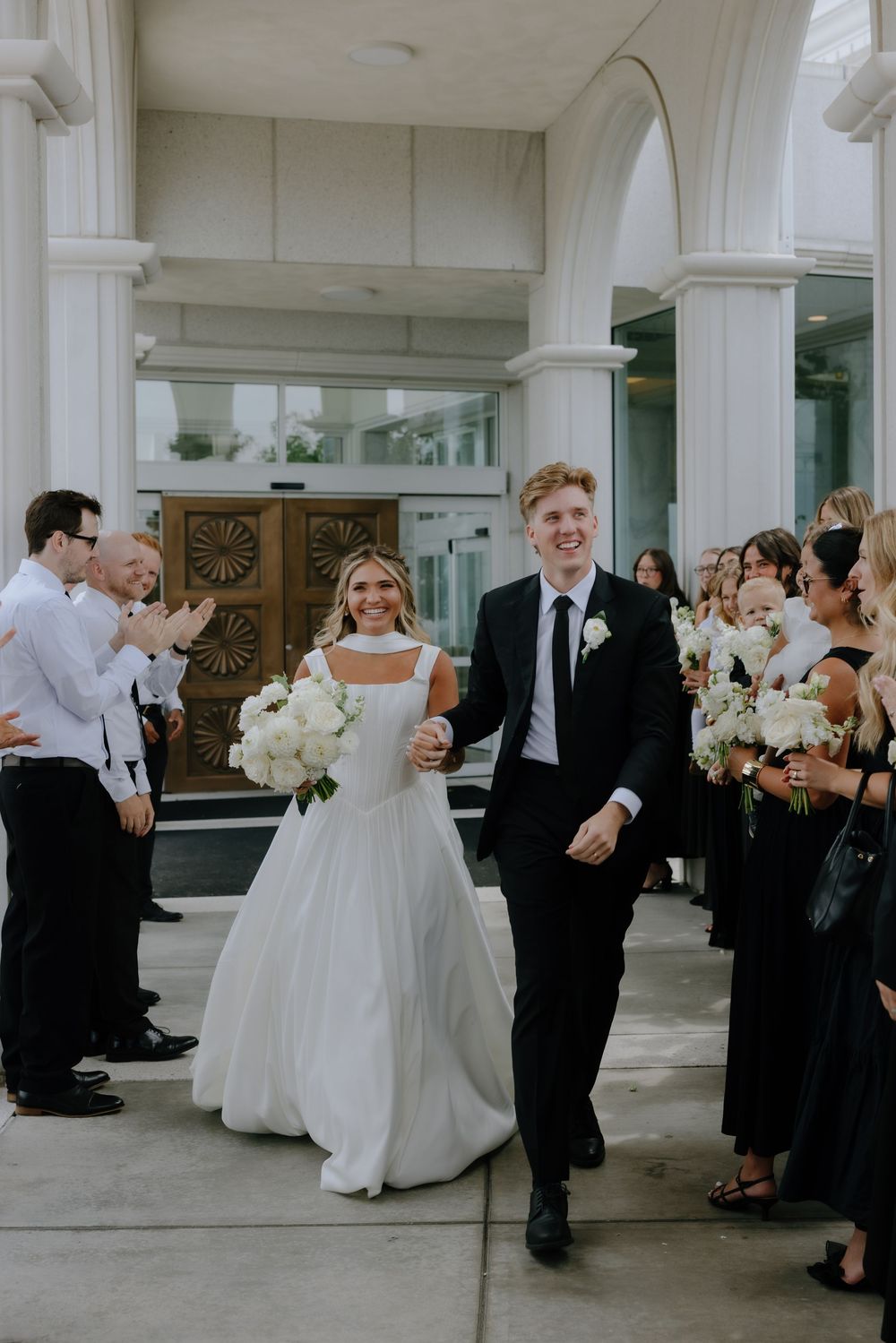 Beaming newlyweds walk through cheering wedding guests holding white bouquets outside a elegant white columned venue.