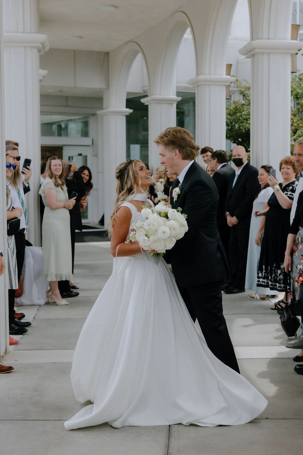 Smiling bride and groom walk through wedding guests under white columns, bride holding white bouquet.