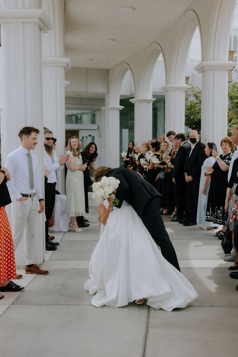 Groom dips bride for romantic kiss outside white columned building as wedding guests cheer.