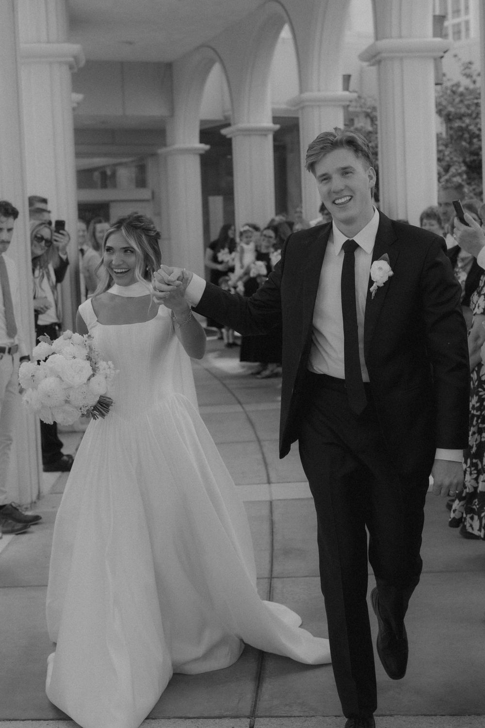 Joyful newlyweds walking down aisle holding hands, bride in white gown carrying bouquet, black and white photo.