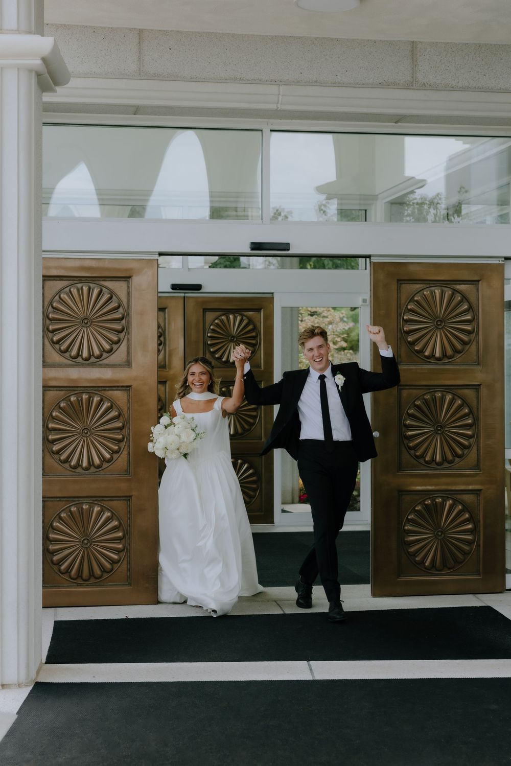 Joyful newlyweds celebrate their exit through ornate wooden doors of a white wedding venue raising hands in triumph.