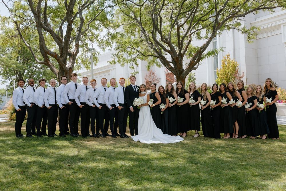 Large wedding party in black and white attire poses on green lawn beneath trees outside a classic white building.