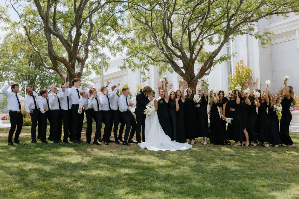 Large wedding party in black attire celebrating with bride and groom under trees on green lawn.