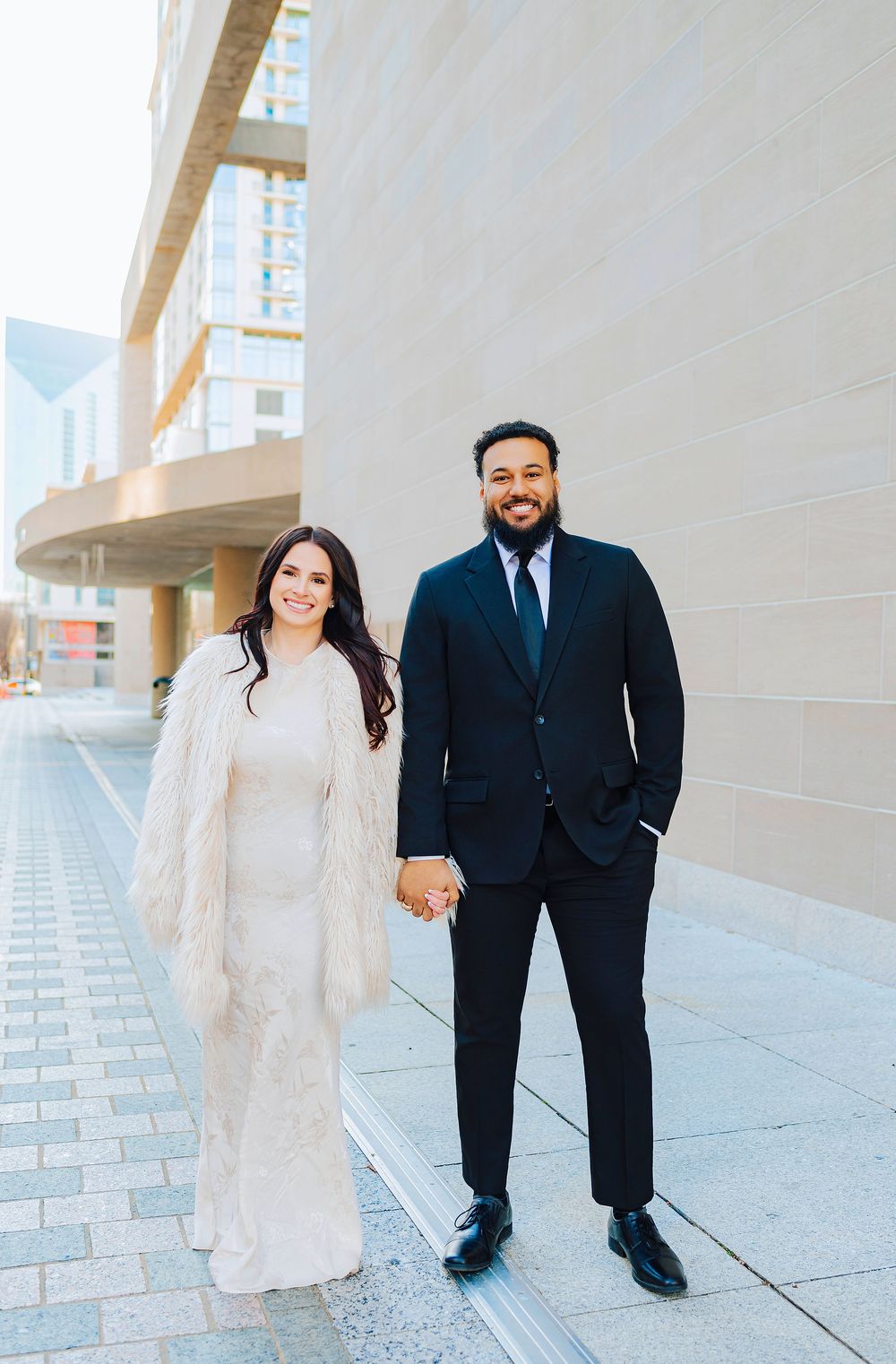Couple posing outdoors in formal attire, woman in white gown with fur coat, man in black suit, holding hands.