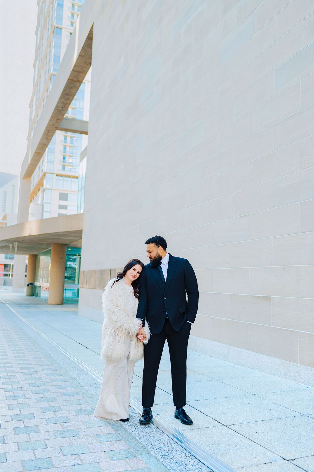 Couple in elegant attire holding hands outside a modern building, woman in white fur coat, man in black suit.