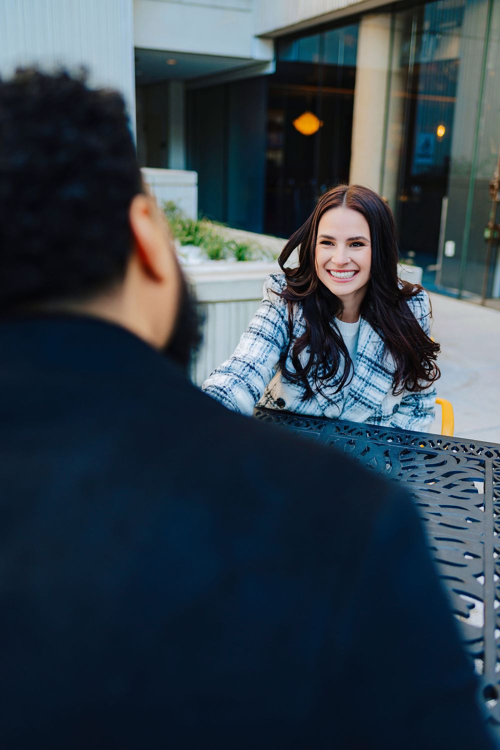 A smiling brunette woman in a plaid jacket sits at an outdoor cafe table, engaged in conversation with a person across from her.