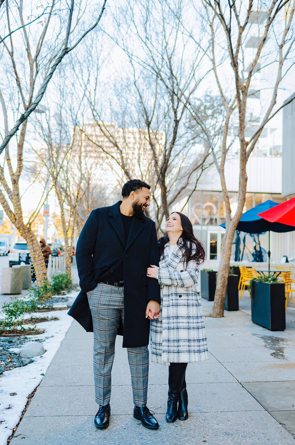 A couple smiles at each other while holding hands on a city sidewalk in winter coats among bare trees.