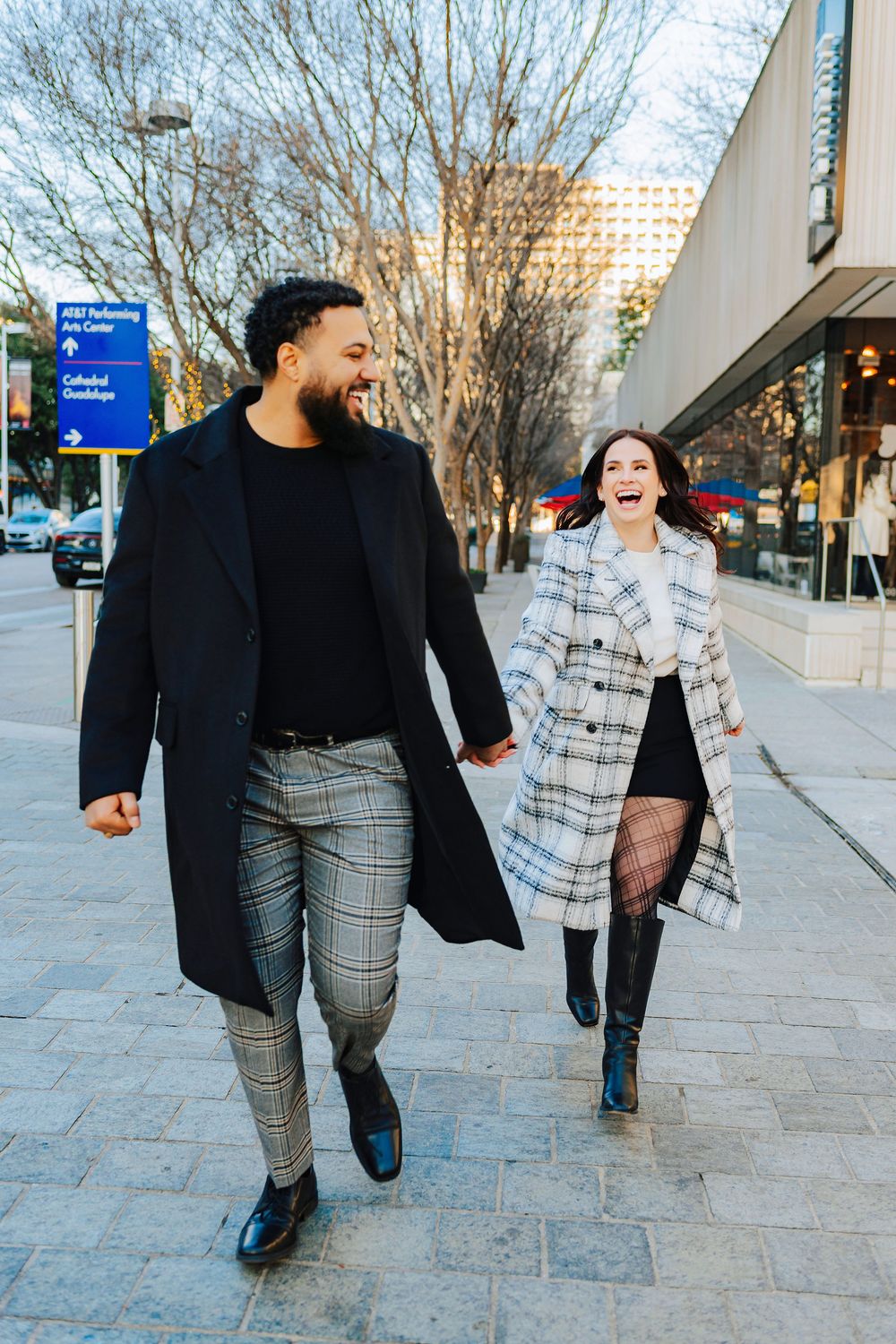 Happy couple laughing and walking hand in hand on city sidewalk wearing coordinating plaid and black coats.
