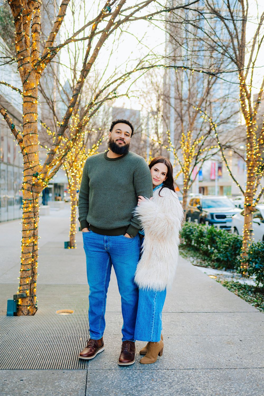 A couple poses on a city sidewalk lined with twinkling fairy lights on bare trees during winter.