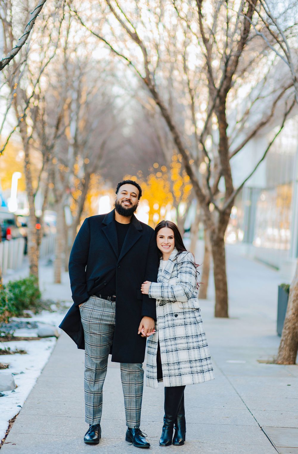 Stylish couple posing on urban sidewalk in coordinating plaid outfits during fall season, smiling at camera.