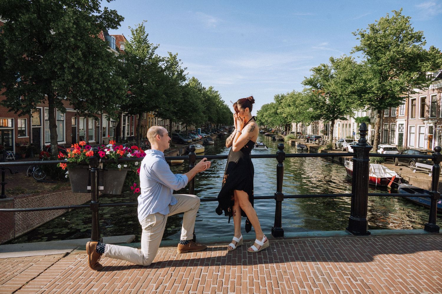 Man in casual clothes kneels on one knee proposing to a woman in a black dress beside a canal railing lined with flowers and trees. Leiden