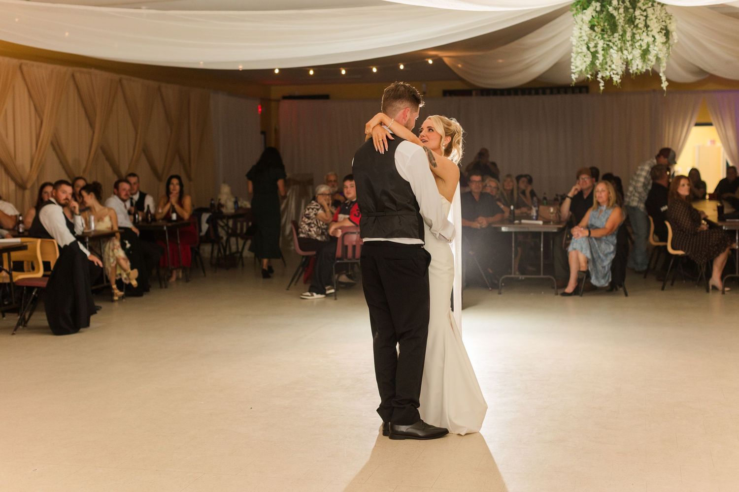 Bride and groom share their first dance on a white dance floor surrounded by wedding guests in a decorated tent venue.