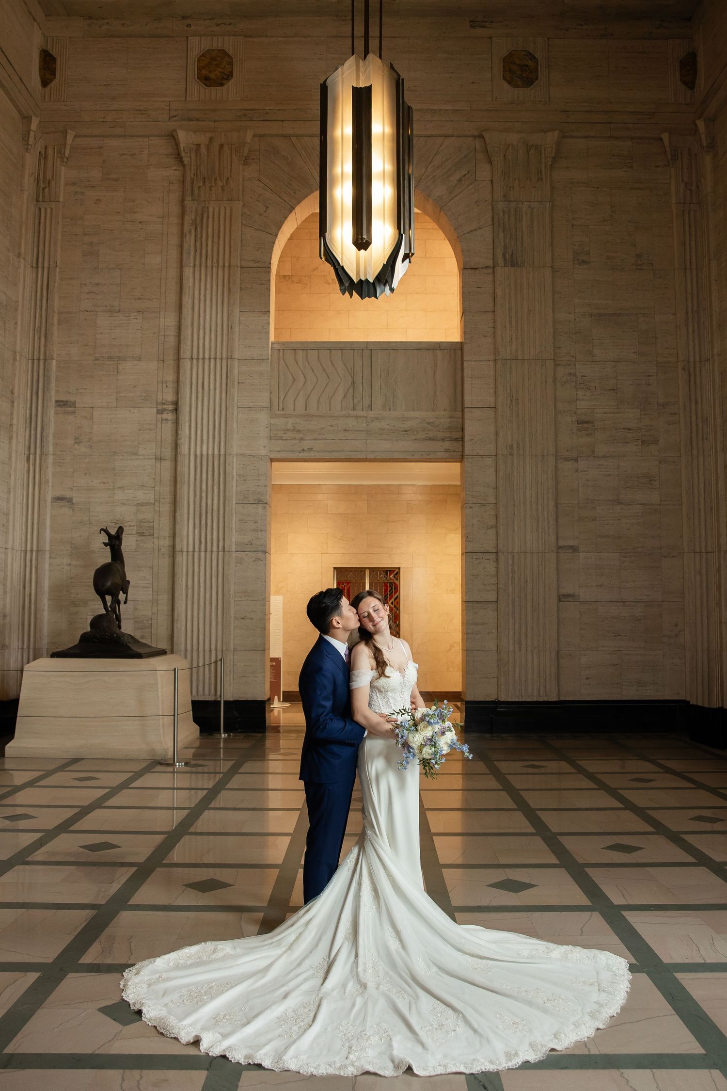 Bride and groom embrace in elegant marble hall, her lace gown trailing beautifully beneath a warm Art Deco chandelier.