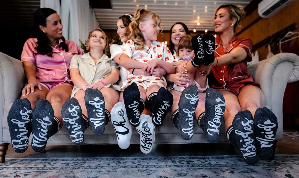 Six women in pajamas sit on a couch showing off custom bridesmaid socks with fun labels like Bride and Maid of Honor.