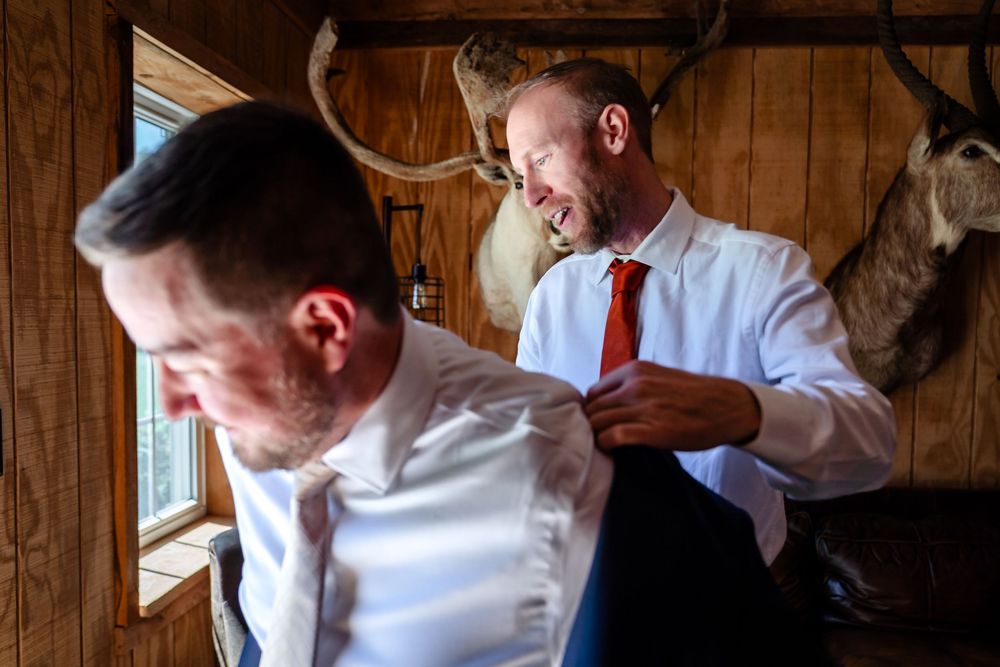 Groomsmen helping each other dress in a rustic cabin with antler decor on wedding day.