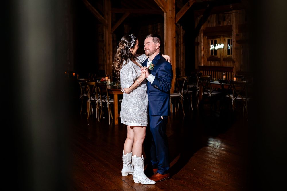 Bride in short silver dress and white boots dances with groom in barn.