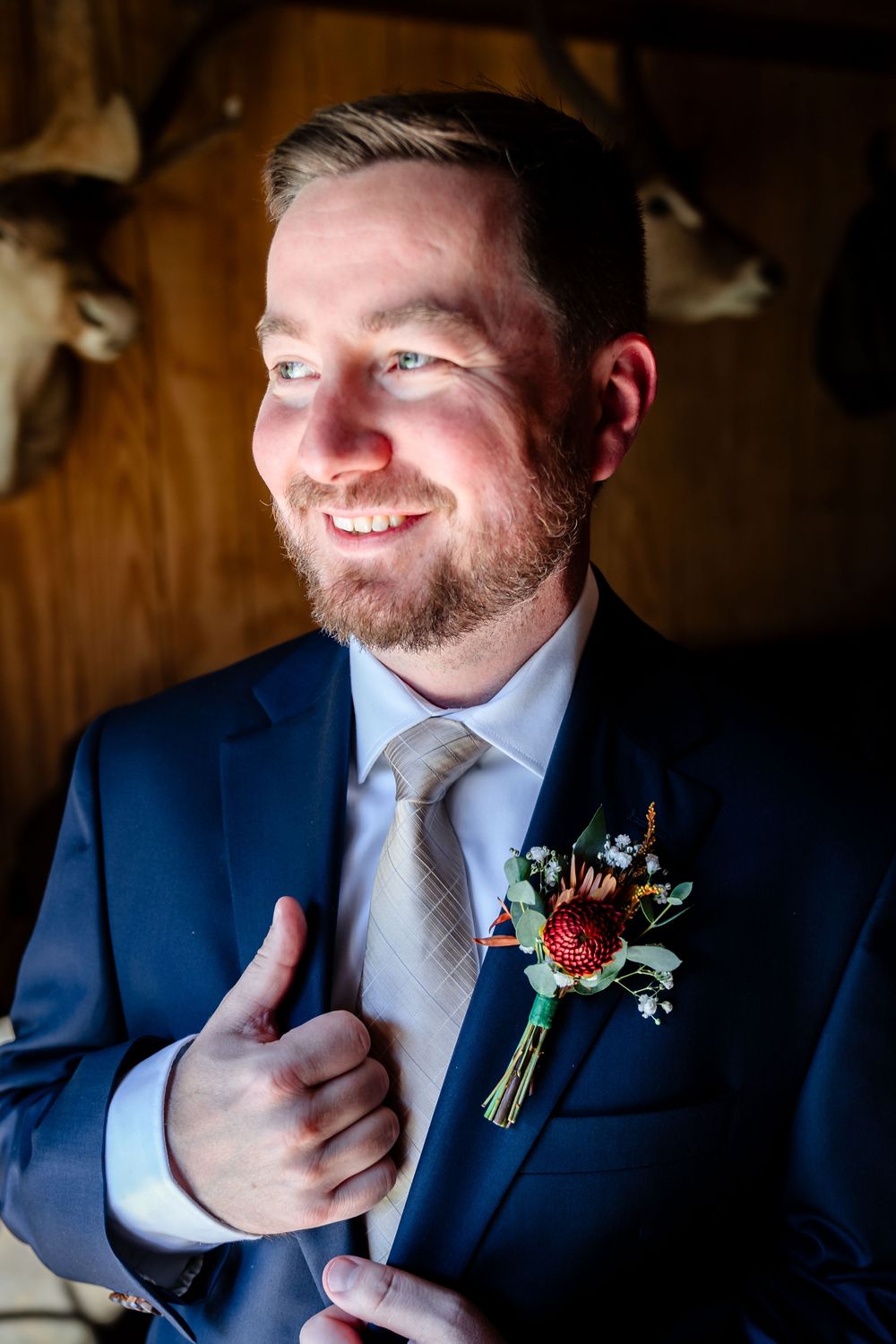 Smiling groom in navy suit with floral boutonniere adjusting jacket, rustic wood background with deer mount.